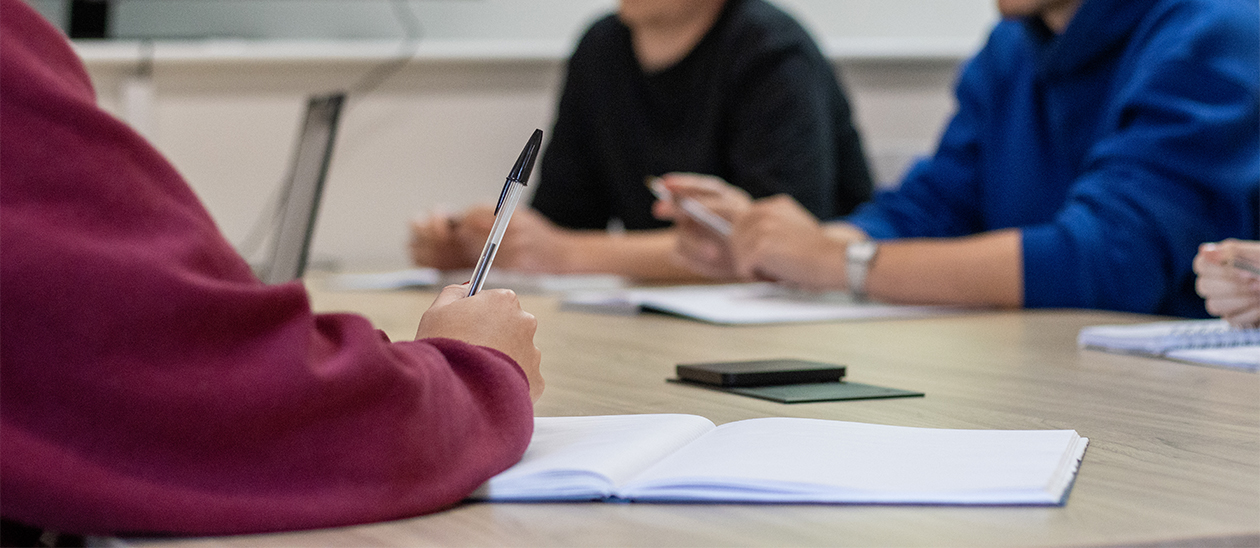 An image of a person taking notes and writing inside a book with two individuals blurred in the background